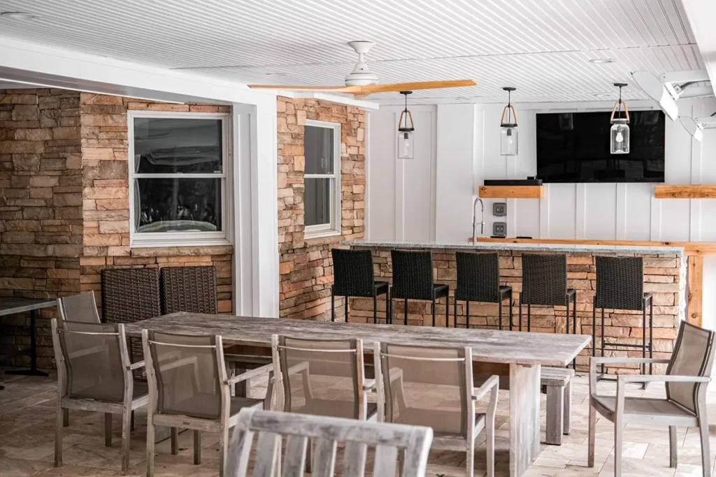 Covered outdoor kitchen and bar under a porch with a beadboard ceiling and stone accent wall